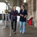 Visually impaired man with white walking aid walking through a station with a woman