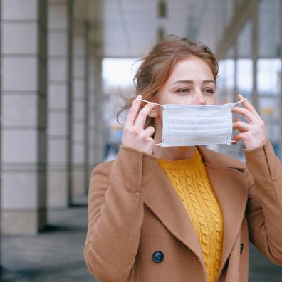 Woman in a brown jacket and yellow jumper placing a disposable face mask on her face