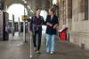 Visually impaired man with white walking aid walking through a station with a woman